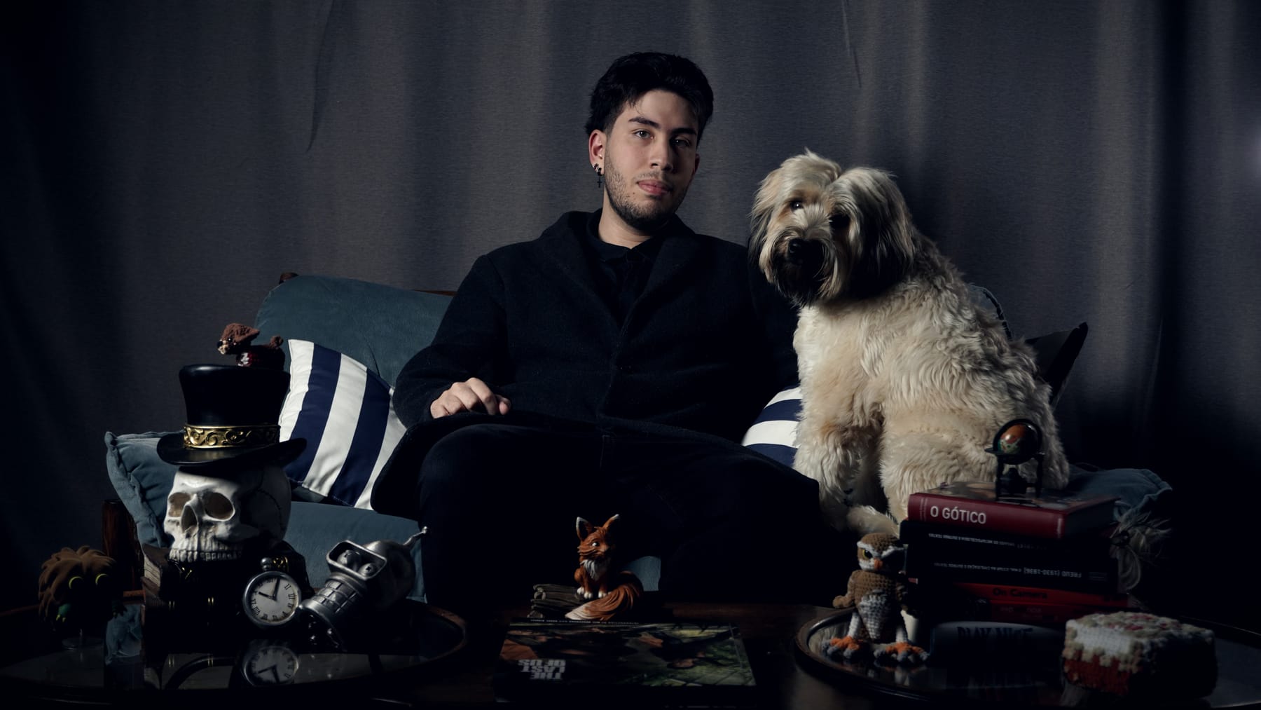 J. V. Dias e seu cão em retrato editorial de estúdio, sentados em um sofá cercados por livros e objetos de cinema e cultura pop